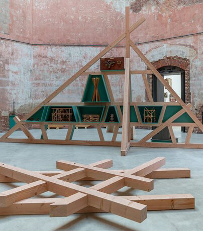 Ein Kunstwerk aus Holzbalken liegt in einem großen Raum mit Ziegelmauerwerk